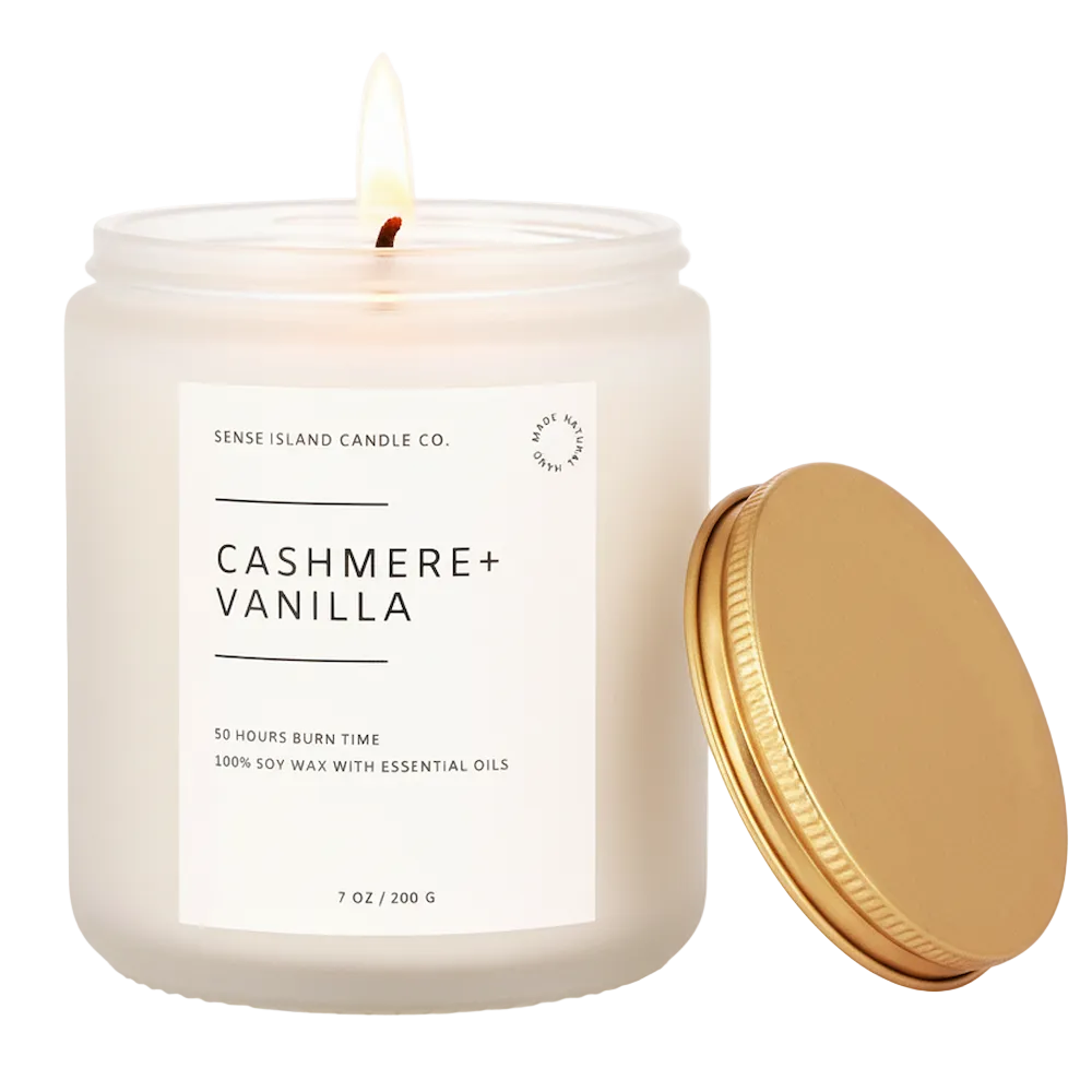 A lit white soy wax candle in a clear glass jar with a white label reading 'CASHMERE + VANILLA' and 'SENSE ISLAND CANDLE CO.' beside a gold metal lid on a white background.